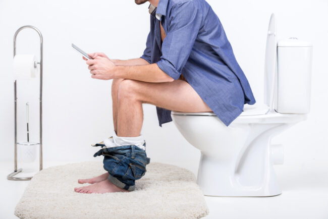 Mann sitzt mit Smartphone auf der Toilette